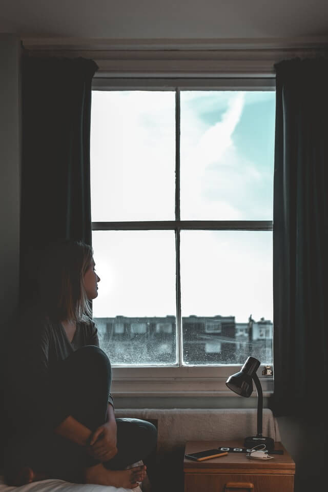 A woman is sitting beside a window looking out from the inside. The window is old and visibly worn. It is a poor insulator. 