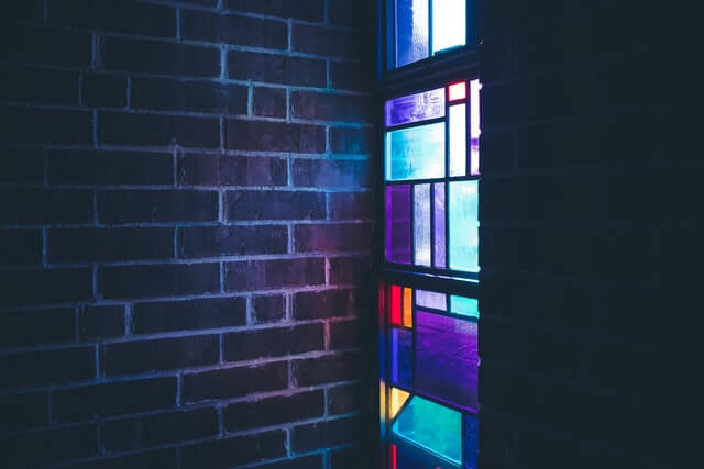 Light shines through a stained glass window into a dark room with vrick walls.