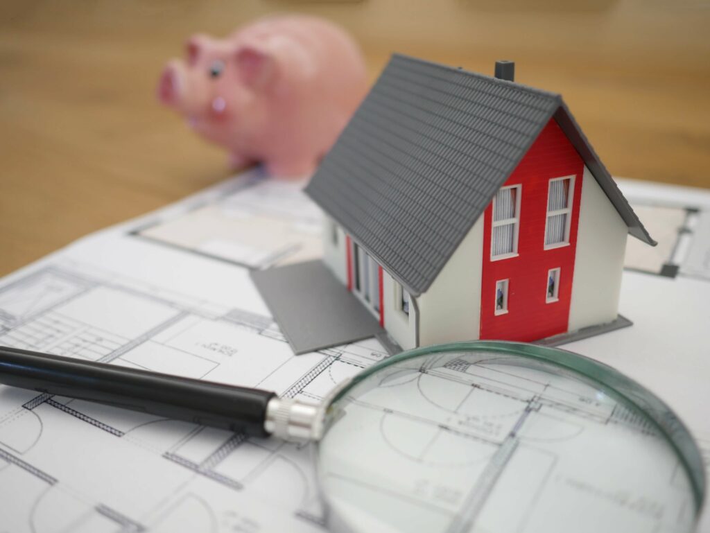 A 3D model of a house rests on a building plan beside a piggy bank and a magnifying glass.