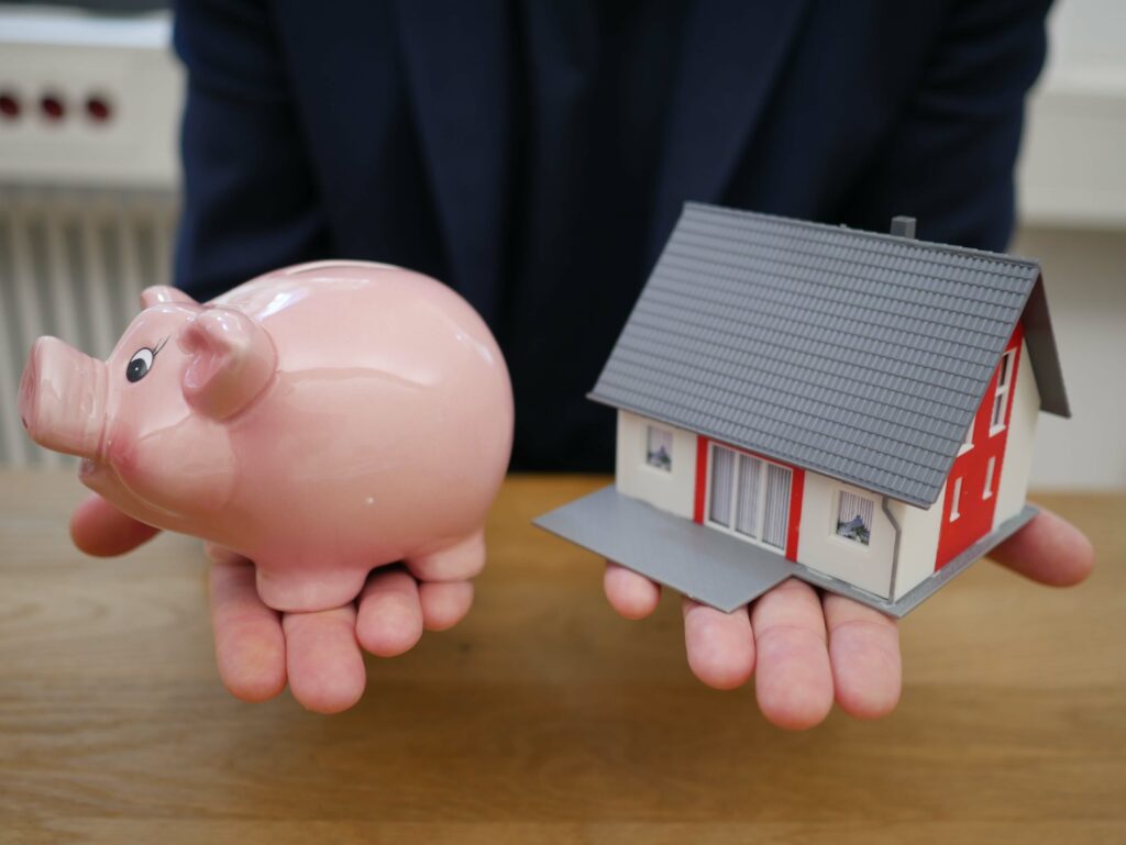 A person holding a piggy bank in one hand and a 3d model house in the other. They are presenting a choice.